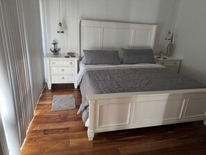 Ein Schlafzimmer mit einem weißen Bett, grauer Bettwäsche, Holzboden, einem weißen Nachttisch und einem kleinen Teppich.