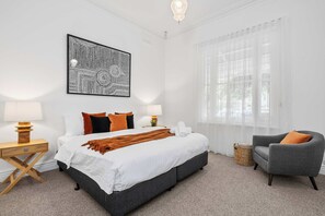 Une chambre d’hôtel avec un grand lit, deux tables de chevet, une chaise, une grande fenêtre avec des rideaux légers et une œuvre d’art murale.