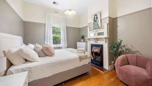 A bedroom with a bed, bedside table, a pink armchair, a fireplace, and a window with curtains. - Character-Filled, Modern Retreat In Mount Pleasant, Ballarat (Mount Pleasant)