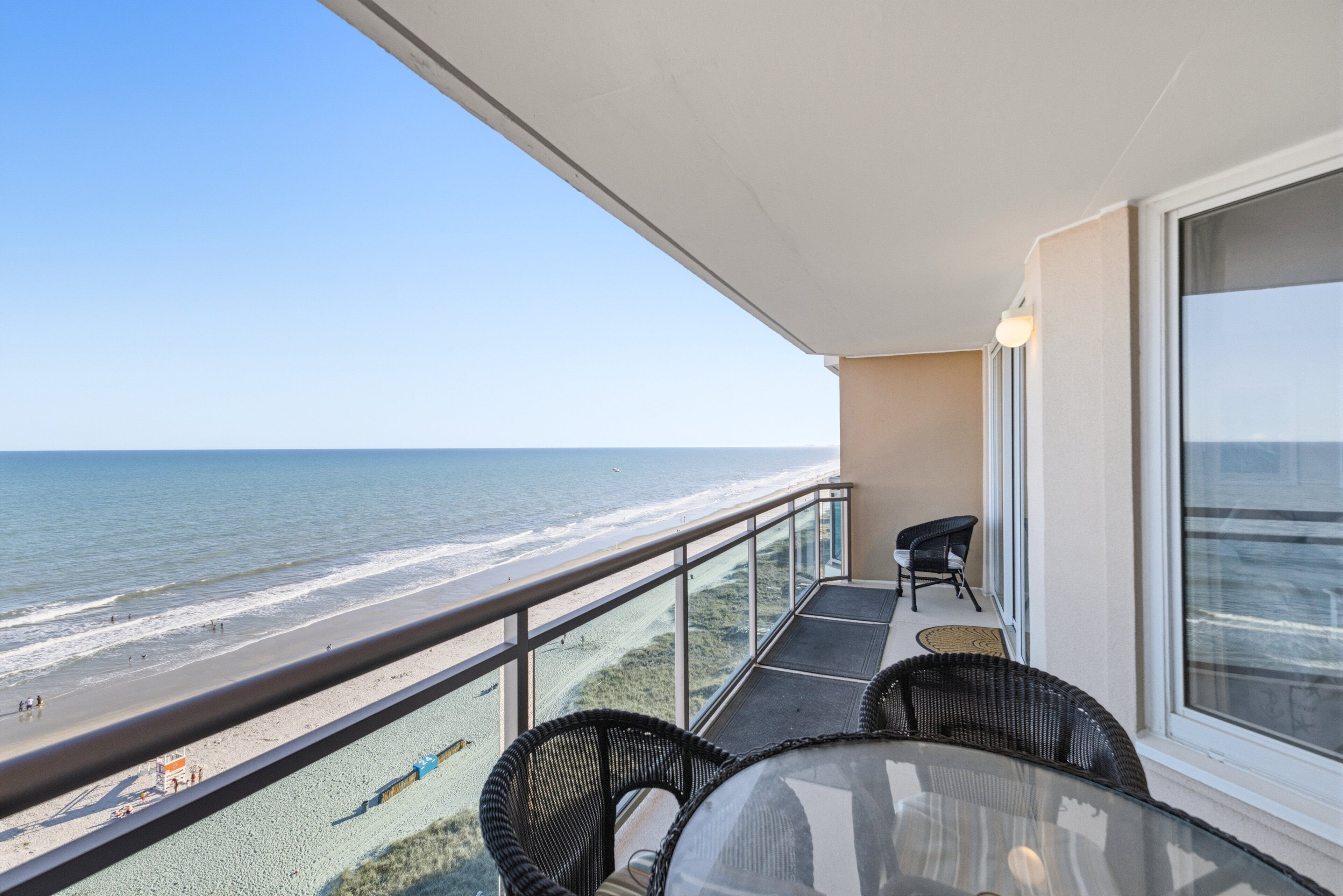 A balcony with a view of the beach, wicker furniture, and a glass table.