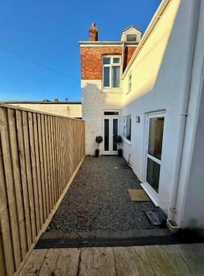 Un patio estrecho con cerca de madera, un sendero de grava y un edificio de fachada blanca y chimenea de ladrillo.