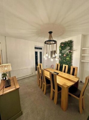 Un comedor con mesa y sillas de madera, una lámpara de araña, un adorno verde en la pared y una lámpara en una mesita auxiliar.