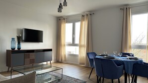 Een moderne woonkamer met een flatscreen televisie, een glazen salontafel, een eethoek met blauwe stoelen en een groot raam met gordijnen.