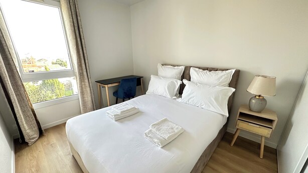 Een opgemaakt bed met witte lakens en kussens, een klein bureau met stoel, een nachtkastje met lamp en een raam met gordijnen.
