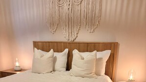 Una cama bien tendida con cabecera de madera, dos mesitas de noche con lámparas y un colgador de macramé en la pared.
