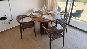 Un comedor con una mesa redonda, sillas de mimbre, una lámpara colgante, un jarrón blanco y un cuadro de una playa.