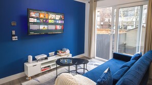 Una sala de estar moderna con una pared azul, un televisor de pantalla plana montado sobre un mueble blanco, un sofá azul y un ventanal corredero que da a un balcón.