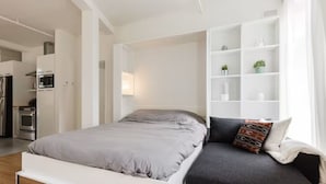 Et moderne soveværelse med seng, sofa, indbygget hvidt reolsystem og et tekøkken i baggrunden.