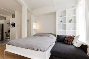 A modern bedroom with a bed, a sofa, a built-in white shelving unit, and a kitchenette in the background. - Apartment in downtown Quebec City (Parking) A (Québec)