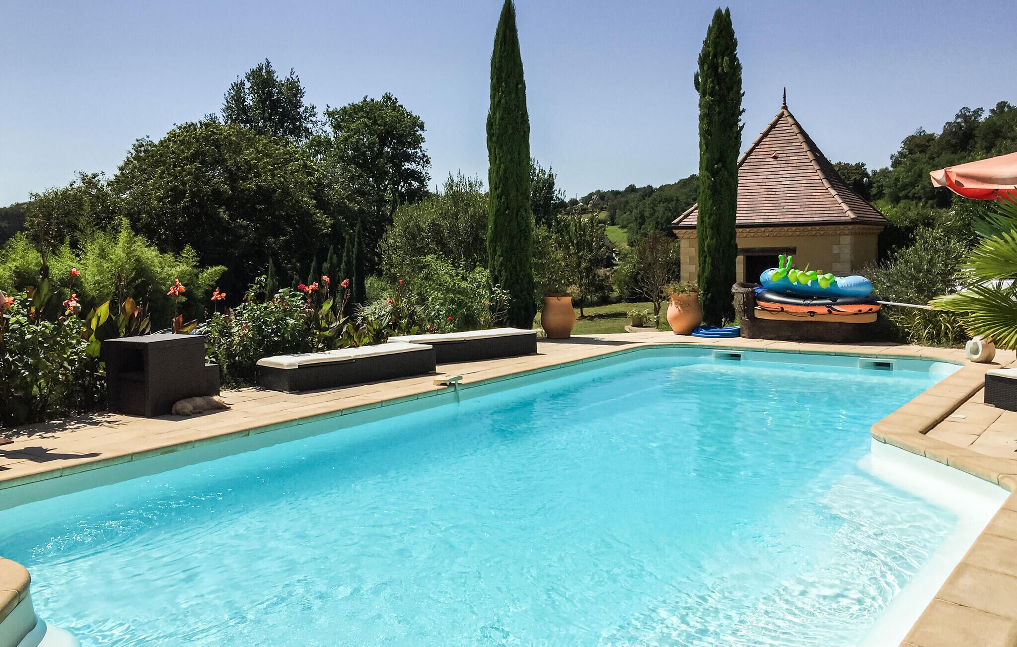 Una piscina amb un solàrium enrajolat, envoltada de vegetació i un edifici petit amb teulada enrajolada.