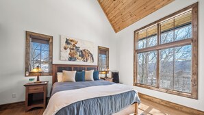 A bedroom with a large bed, wooden headboard, two bedside tables with lamps, a large window with a view, and a painting on the wall. - Sundance Lodge at Eagles Nest - hot tub, mtn home, club amenities (Elk Park)