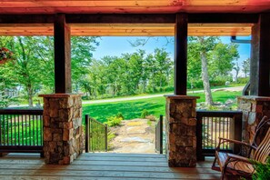 A wooden porch with stone pillars, a bench, and a view of a green landscape with trees and a road. - The Sugar Chalet - Luxury home on Sugar, easy access, views! (Sugar Mountain)