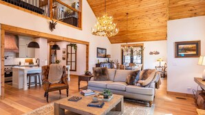 A spacious living area with a high ceiling, a chandelier, a sofa, a coffee table, and a kitchen in the background. - The Sugar Chalet - Luxury home on Sugar, easy access, views! (Sugar Mountain)