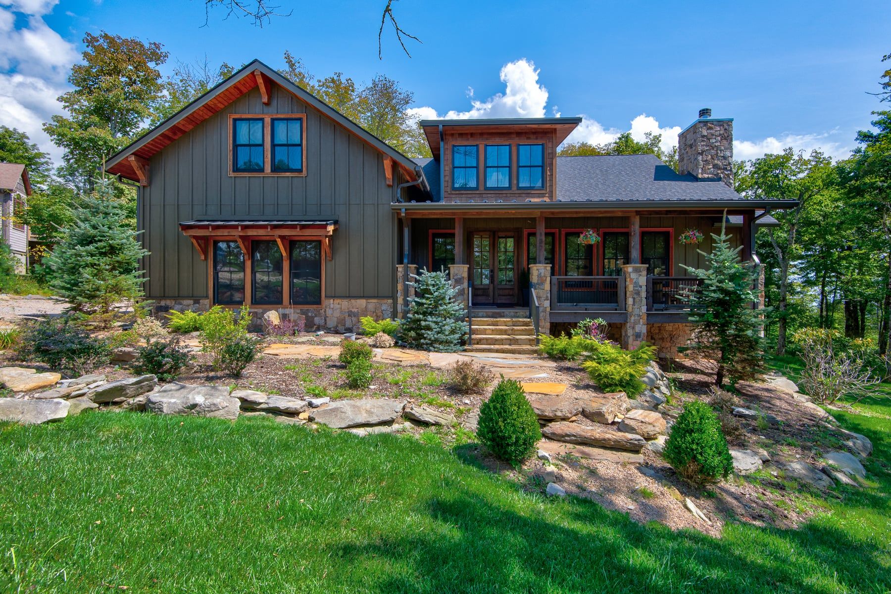 A two-story house with a stone chimney, a porch with stone pillars, and a well-maintained garden with rocks and shrubs.