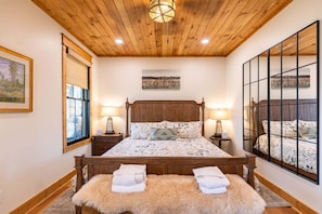 A bedroom with a wooden ceiling, a large bed with floral bedding, two bedside tables with lamps, and a large window with a view. - The Sugar Chalet - Luxury home on Sugar, easy access, views! (Sugar Mountain)