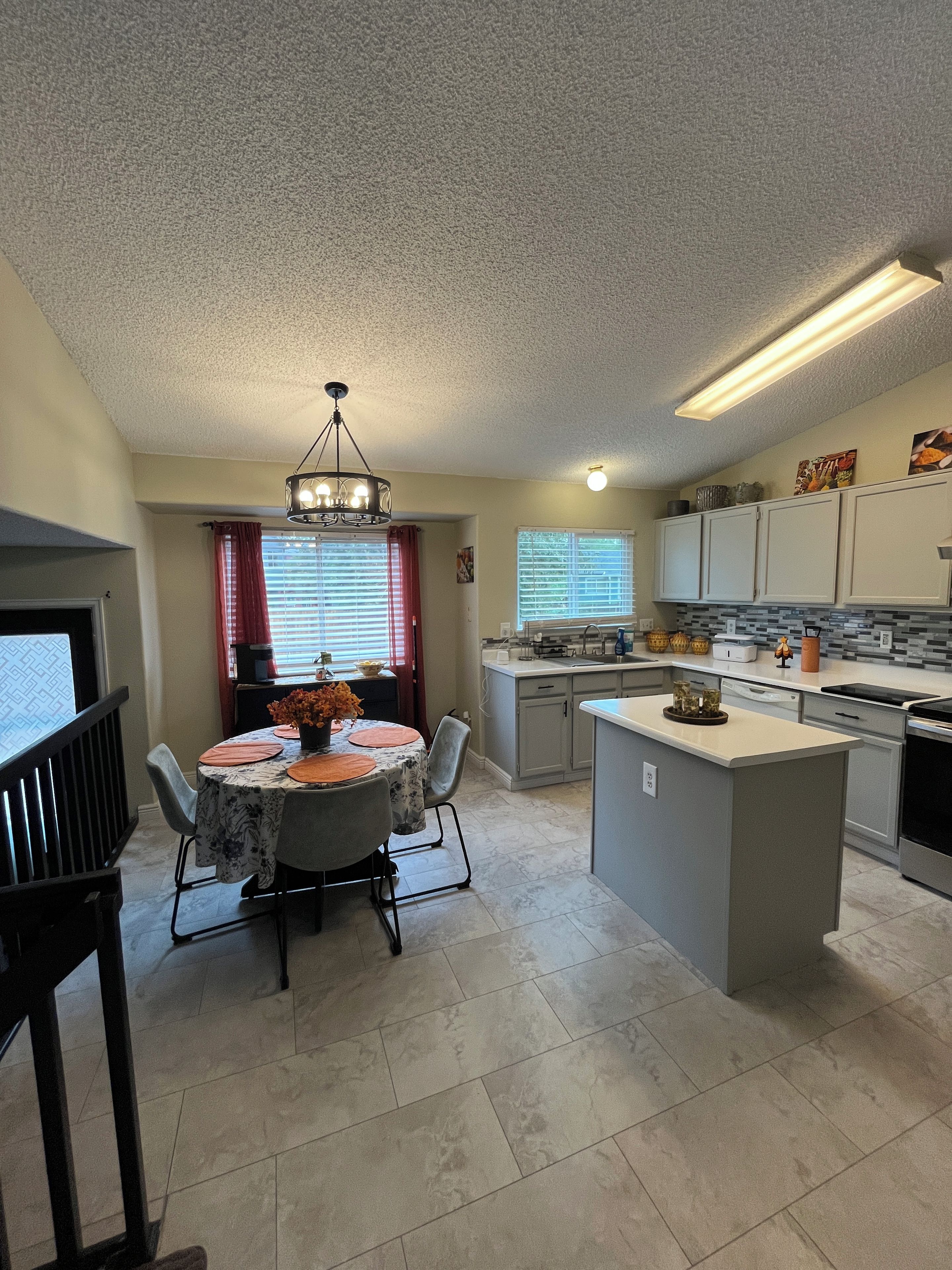 A modern kitchen with a central island, a dining area with a chandelier, and a view of the outside through a window with blinds.