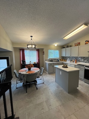 A modern kitchen with a central island, a dining area with a chandelier, and a view of the outside through a window with blinds. - Spacious DIA House 3BR + Bonus Bed House (Denver)