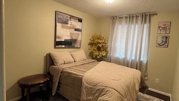 Een slaapkamer met een bed, een tafeltje, een vloerkleed, een plant en een schilderij aan de muur.