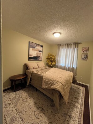A bedroom with a bed, a small table, a rug, a plant, and a painting on the wall. - Spacious DIA House 3BR + Bonus Bed House (Denver)