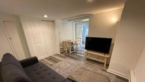 A living room with a sofa, a TV on a stand, a dining table with chairs, and a door. - 1740 Le Cocon Limoilou Enfant-Jésus Hospital (Québec)