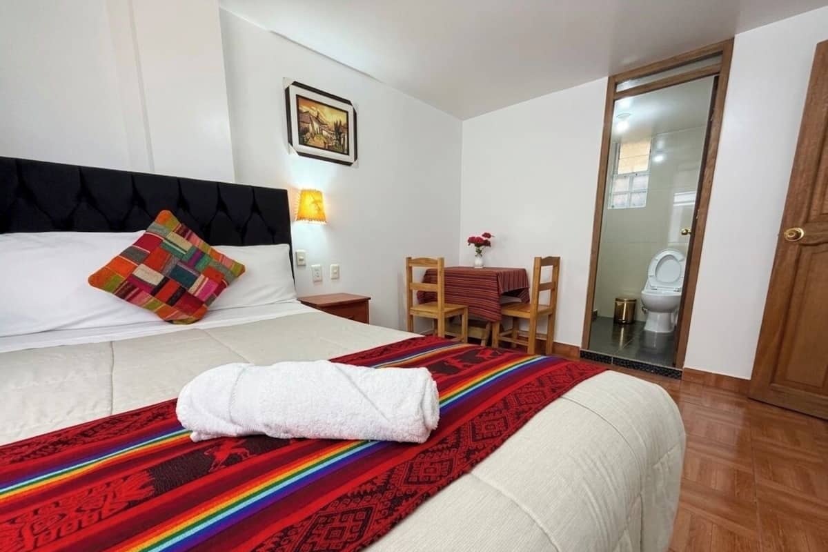 Hotellihuone, jossa on sänky, yöpöytä lampun kanssa, pieni pöytä maljakolla ja ovi, joka johtaa kylpyhuoneeseen.