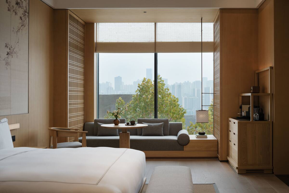 A modern hotel room with a large bed, a sofa, a small table with a vase, and a view of the cityscape through large windows.