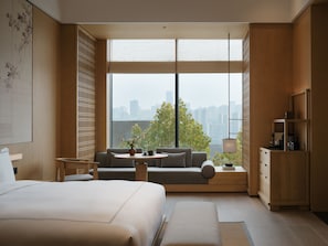 Une chambre d’hôtel moderne avec un grand lit, un canapé, une petite table avec un vase, et une vue sur le paysage urbain grâce à de grandes fenêtres.
