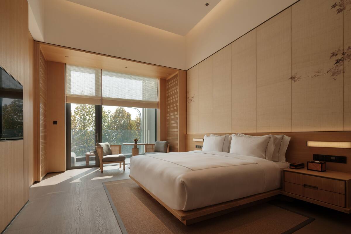 A modern bedroom with a large bed, a bedside table, a chair, and a window with a view.