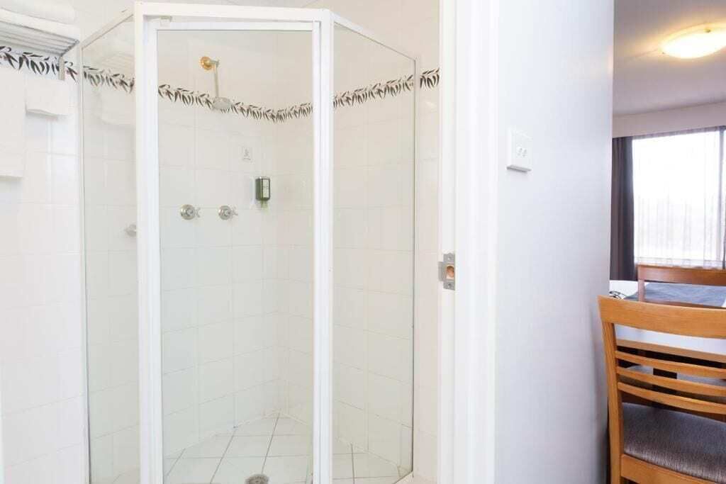 A glass-enclosed shower with a white tiled wall, a wooden chair, and a window with curtains.