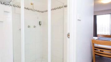 A glass-enclosed shower with a white tiled wall, a wooden chair, and a window with curtains.