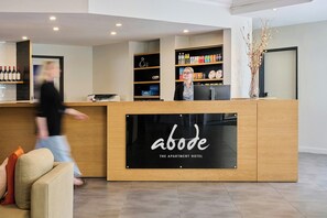 A reception desk with a sign reading 'abode THE APARTMENT HOTEL,' a counter with wine bottles, and a person walking past. - Alpha Hotel Canberra (Canberra)