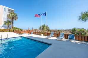 Una piscina con sillas de descanso y una valla de madera, un asta con dos banderas, y un edificio con balcones.