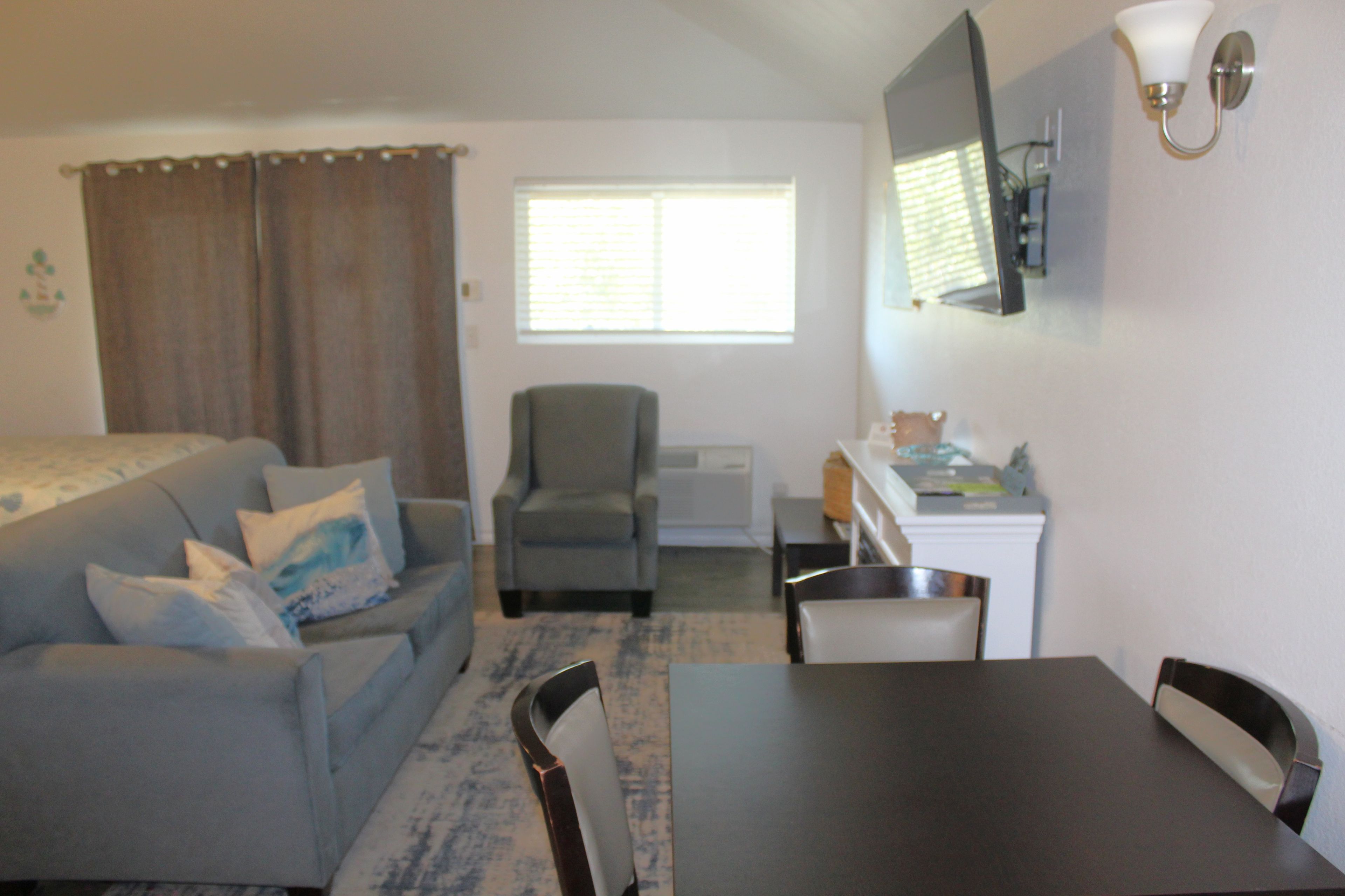 A small, furnished room with a sofa, a dining table, a chair, a television mounted on the wall, and a window with blinds.