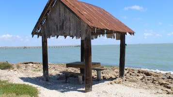 A wooden structure with a bench and table, located on a pebbled beach with a body of water in the background.