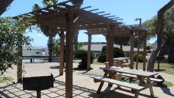 Una estructura de pergola de fusta amb taules de pícnic i bancs adossats, envoltada d'una zona pavimentada amb palmeres i vista a l'oceà.