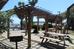 Una estructura de pergola de fusta amb taules de pícnic i bancs adossats, envoltada d'una zona pavimentada amb palmeres i vista a l'oceà.