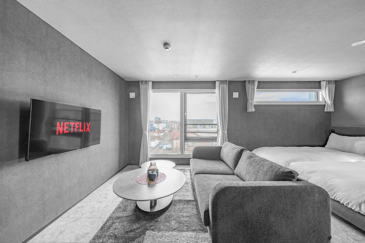 Ein modernes Hotelzimmer mit großem Fernseher, der 'NETFLIX' anzeigt, einer Couch, einem Bett, einem kleinen Tisch mit Vase und Blick auf die Stadt durch große Fenster.