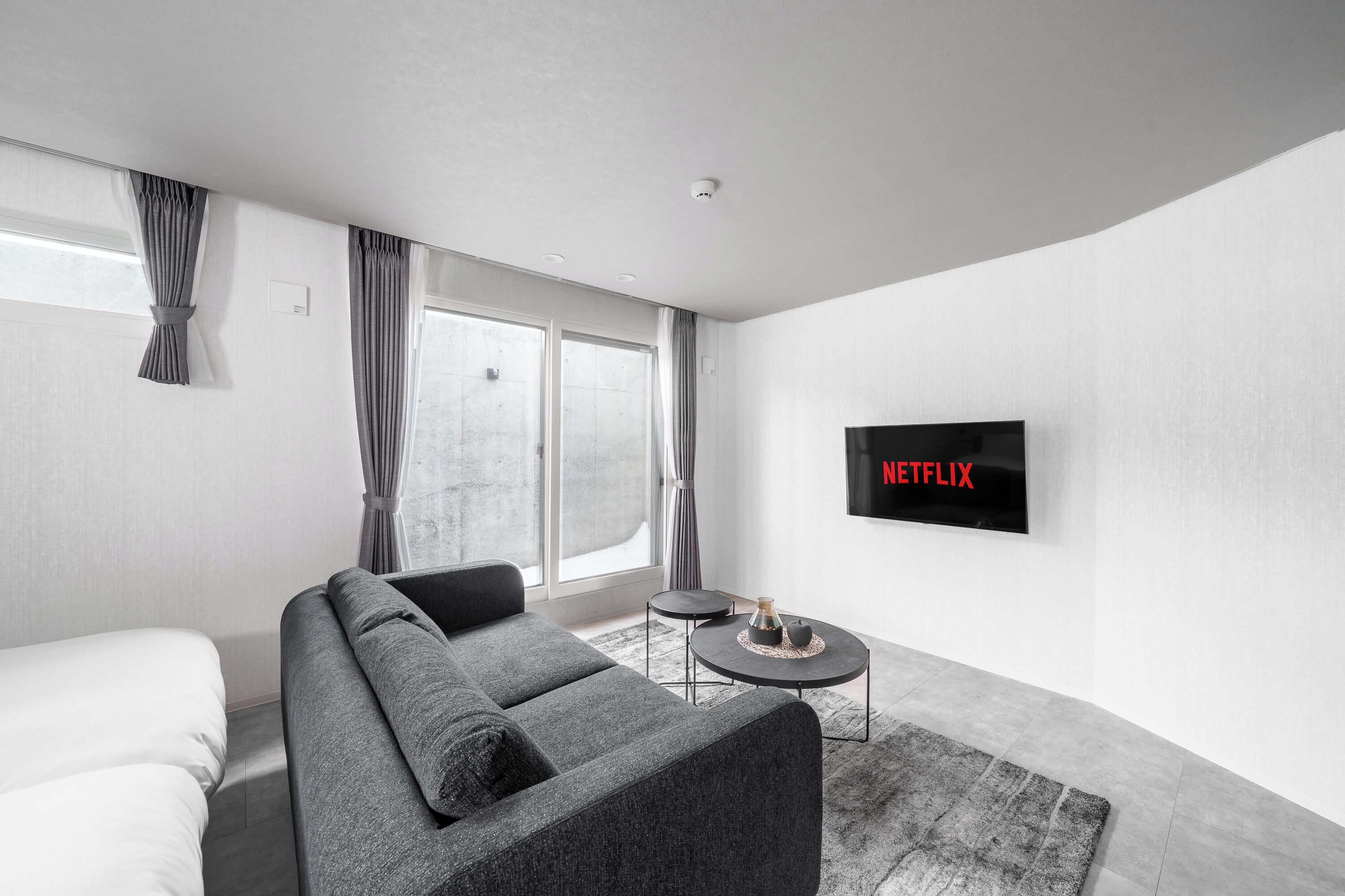 A modern living room with a grey sofa, a small round table, a flat-screen TV mounted on the wall, and a large window with curtains.