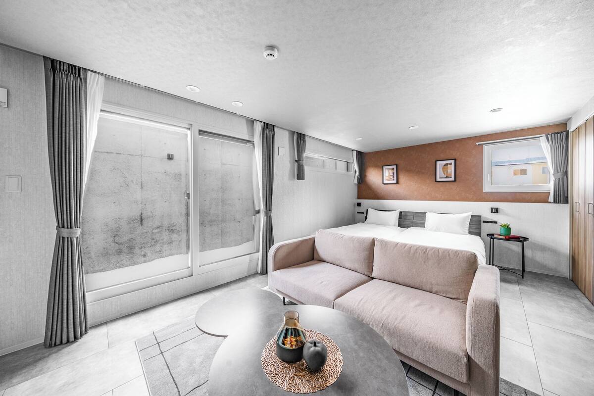 Ein modernes Hotelzimmer mit Schlafsofa, einem kleinen Tisch mit Flasche und Glas, einem Fenster mit Vorhängen und Blick ins Freie.