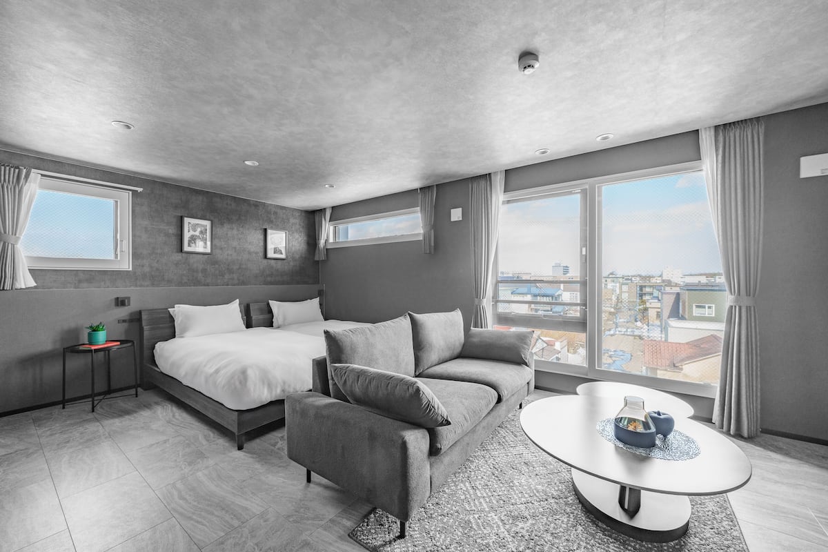Ein modernes Hotelzimmer mit Bett, Sofa und rundem Couchtisch, großen Fenstern mit Vorhängen und Blick auf die Stadt.
