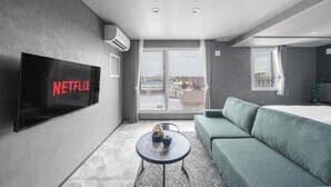 A modern hotel room with a large flat-screen TV, a sofa, a small round table, and a balcony with a view. - GRAN HILLS (Chitose)