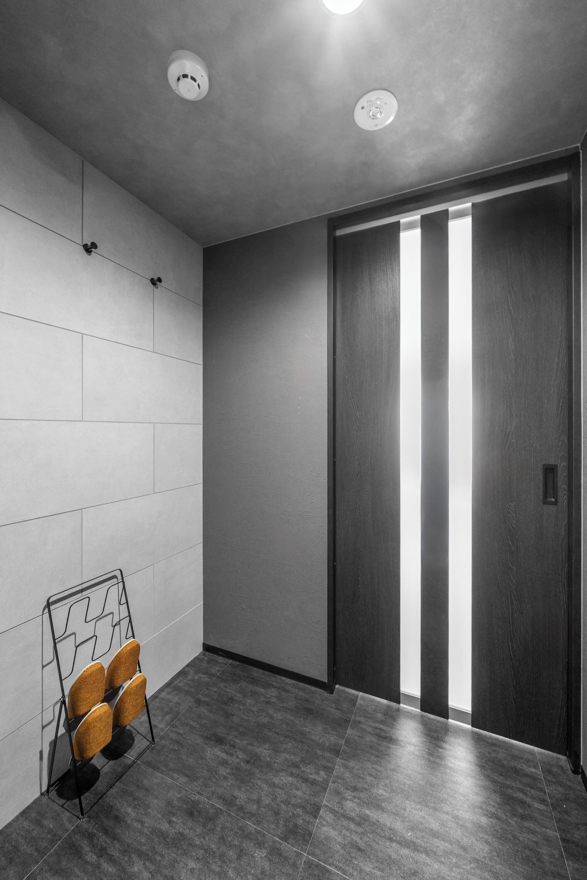Un ascensor moderno con revestimiento oscuro, puerta de vidrio, lámpara de pared y un asidero para sillas con cuatro sillas.