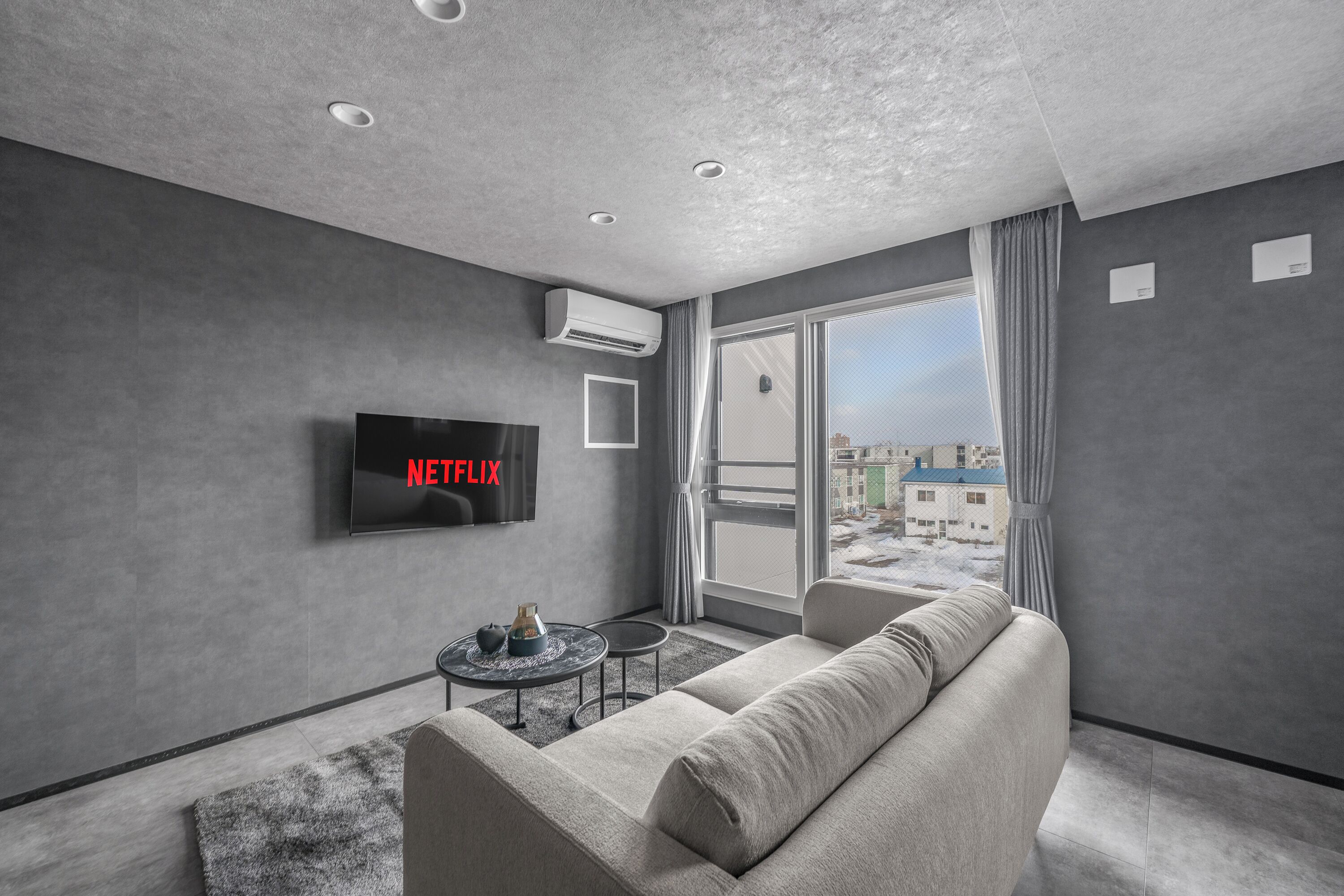 A modern living room with a grey sofa, a flat-screen TV, a small coffee table, and a view of buildings through a large window.