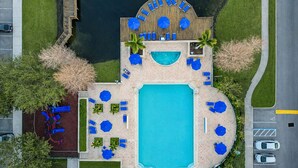Eine Vogelperspektive auf einen Poolbereich mit Liegestühlen und Sonnenschirmen, umgeben von viel Grün.