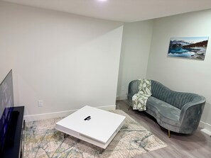 A modern living room with a white coffee table, a grey sofa with a floral throw, a mounted painting of a mountain landscape, and a flat-screen TV.