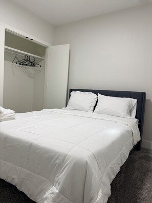 A neatly made bed with white bedding, a dark headboard, and a closet with hanging clothes.