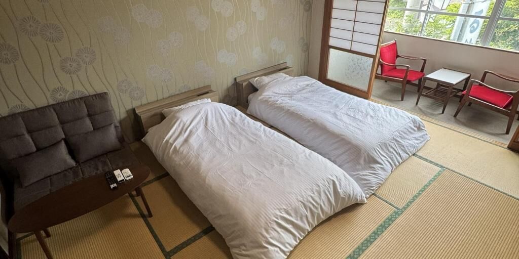 A room with two beds, a sofa, a small table, and a view of greenery through a window.