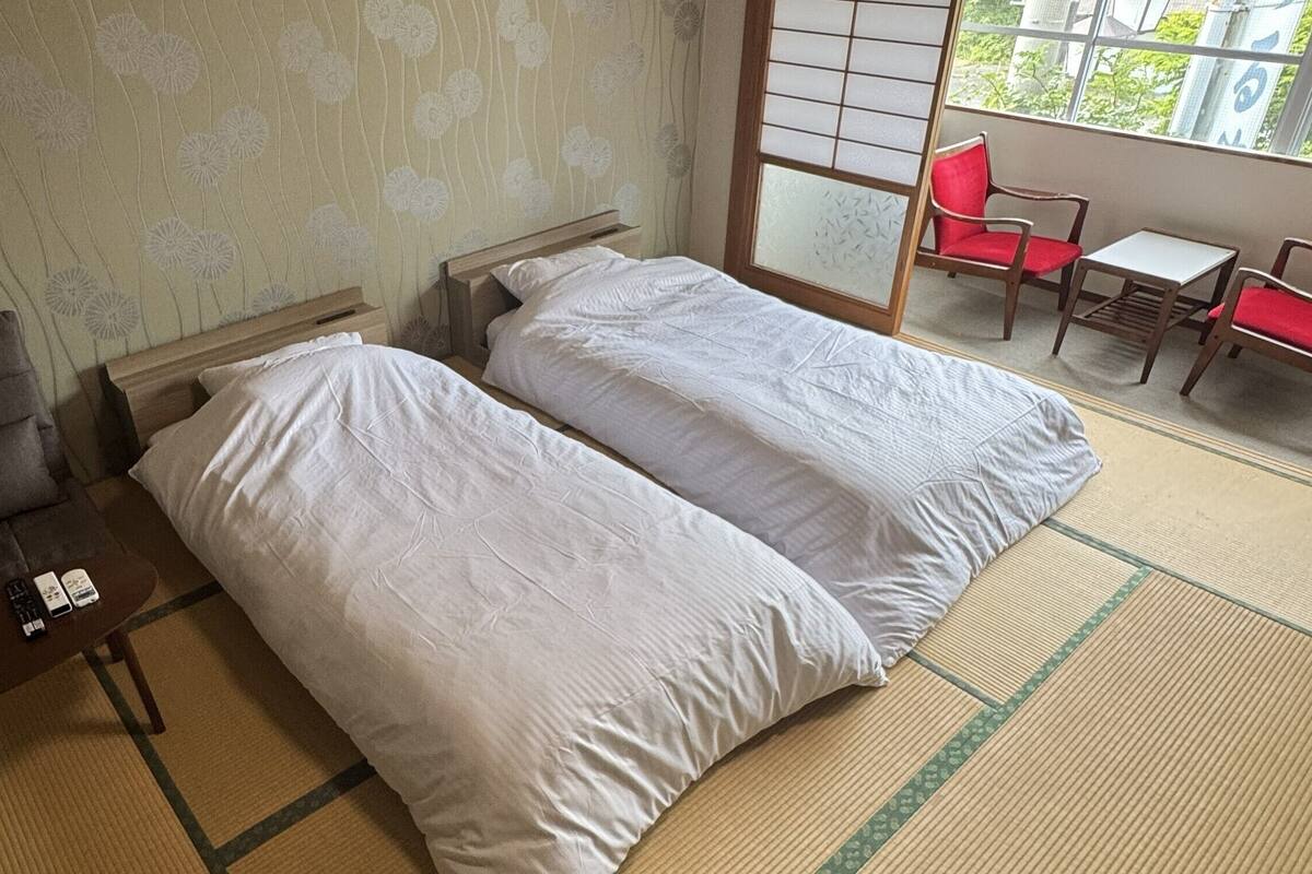 A room with two beds, a sofa, a small table with remote controls, and a view of a balcony with red chairs.