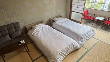 A room with two beds, a sofa, a small table with remote controls, and a view of a balcony with red chairs.
