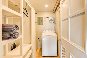 A laundry room with a washing machine, shelves with folded clothes, and a wall-mounted electrical outlet. - Udacha this is a fully renovated studio (Montego Bay)